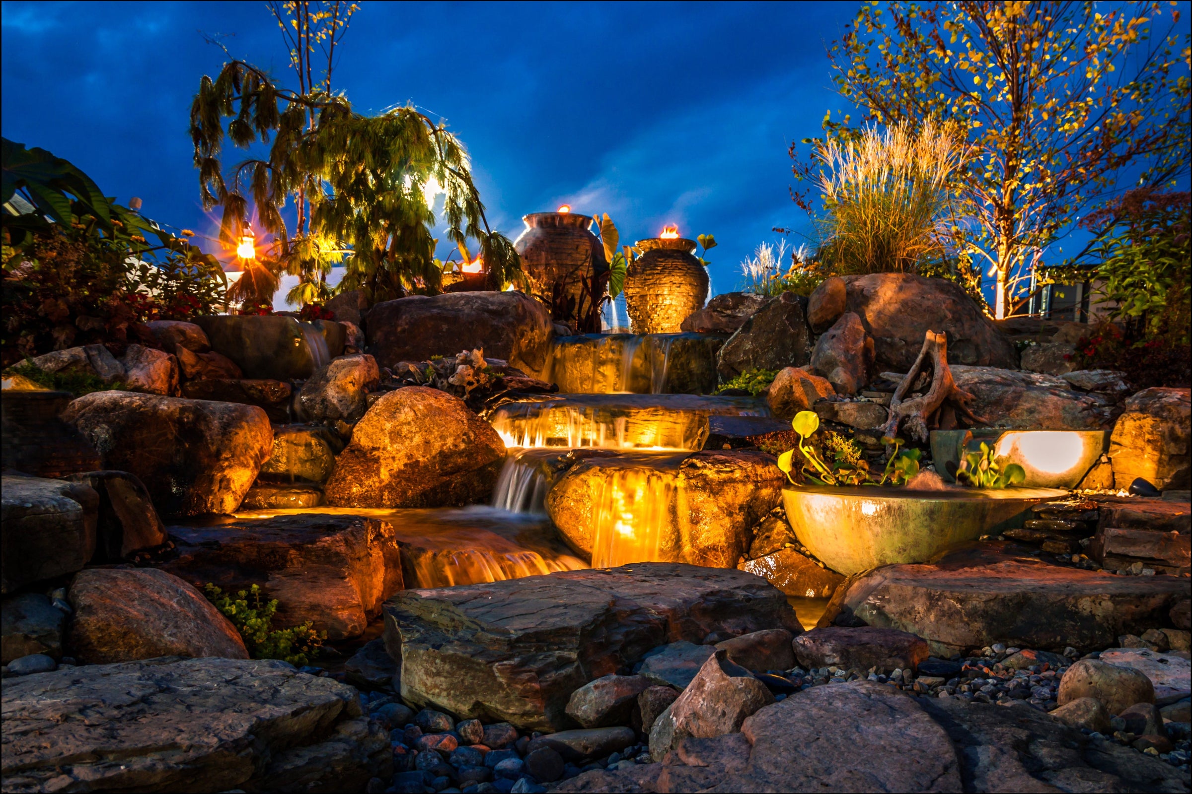 An illuminated outdoor rock waterfall garden feature at dusk, with warm golden lights under a deep blue evening sky. Large natural stones form multiple tiers of flowing water, with decorative ceramic pots, plants, and small trees around the feature.