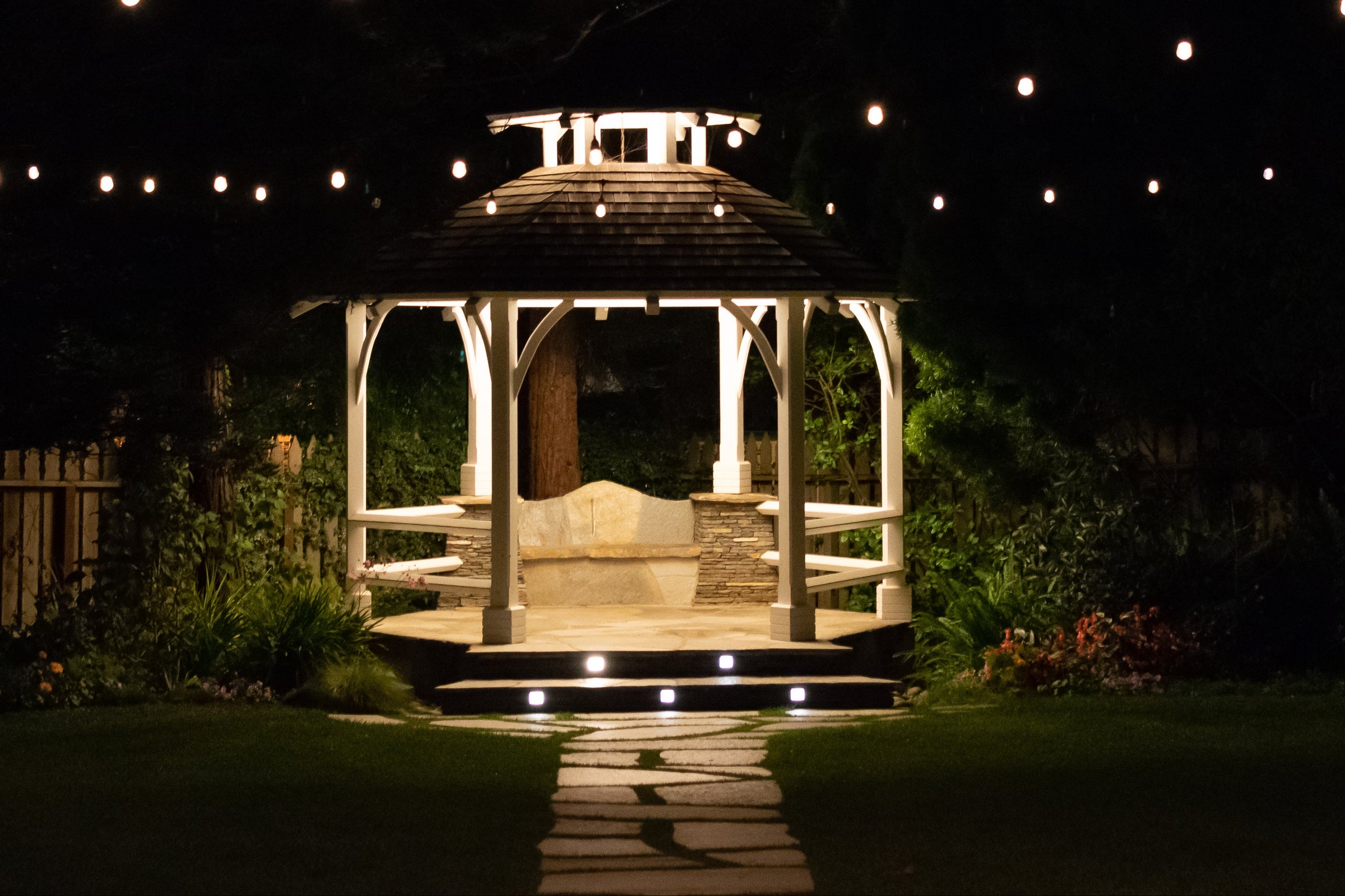 An illuminated white wooden gazebo with a dark shingled roof stands on a stone path at night in a landscaped garden. Warm string lights hang above, and the gazebo is lit from within, creating a cozy outdoor atmosphere.