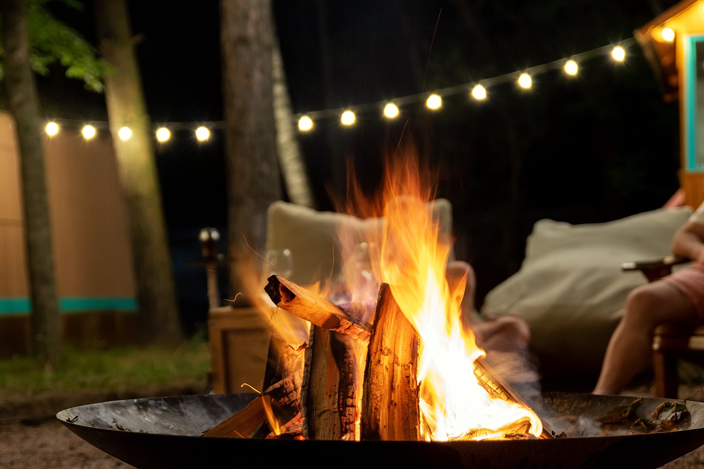 A black metal fire pit bowl containing a bright burning wood log, set outdoors at night with a campsite scene in the background featuring a person sitting in a chair under warm string lights.