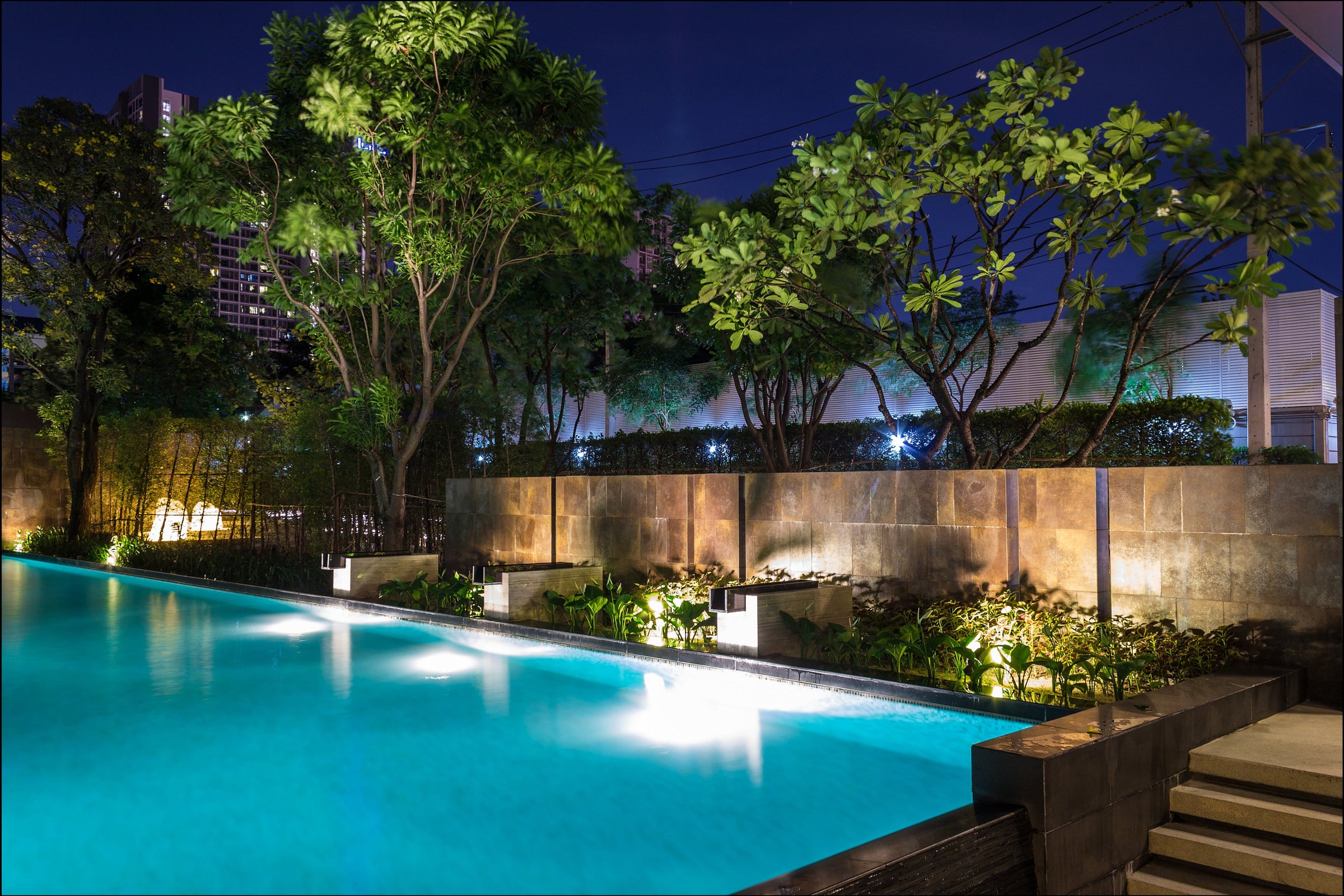 A modern outdoor swimming pool at night with glowing turquoise water, surrounded by a stone wall, illuminated greenery, and trees under a dark night sky.